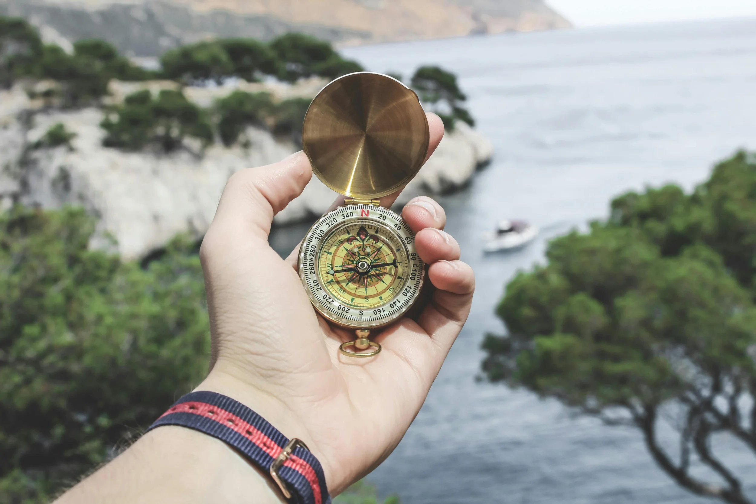 A hand holding a vintage brass compass with a compass rose, pointing towards a scenic coastal landscape with water, trees, and a boat in the background.