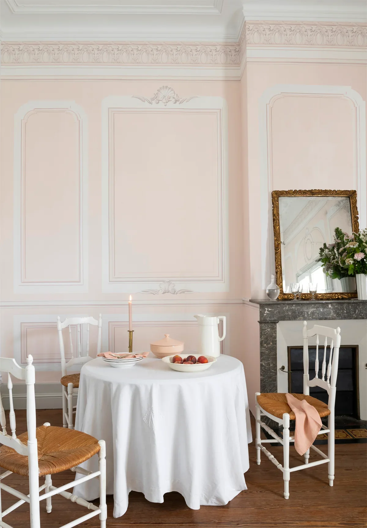 A breakfast table set with a white tablecloth, a pink candle, a bowl of peaches, a pink lidded bowl, a white pitcher, and a plate with pink napkins. Four white chairs with woven seats surround the table. In the background, a mirror with a gold frame 