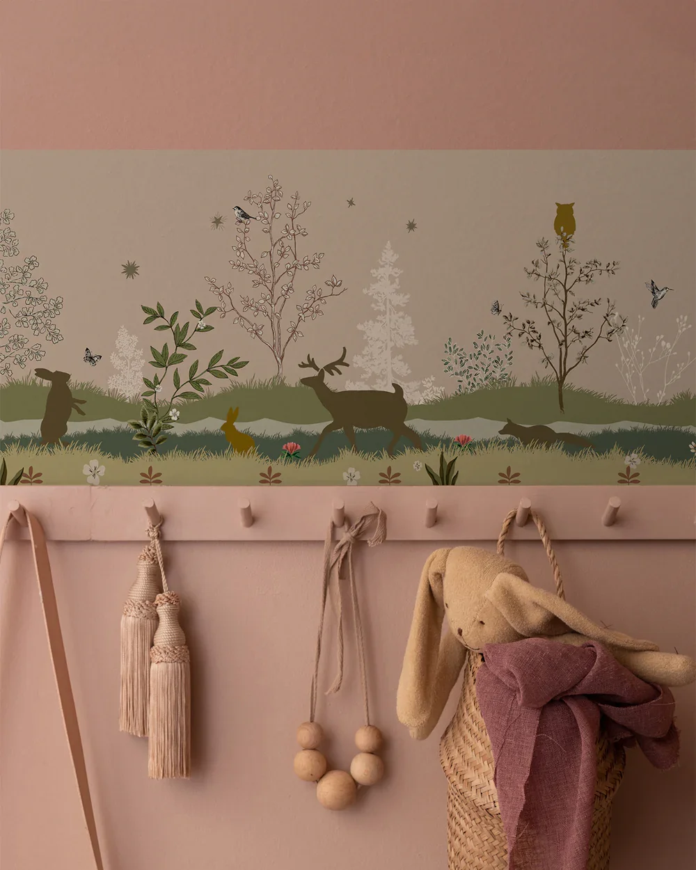 A wall decor mural showing silhouette animals, trees, and butterflies in a forest scene; below, a pink wall with hooks holding tassels, a bead necklace, and a woven basket containing a plush bunny with pink clothes.