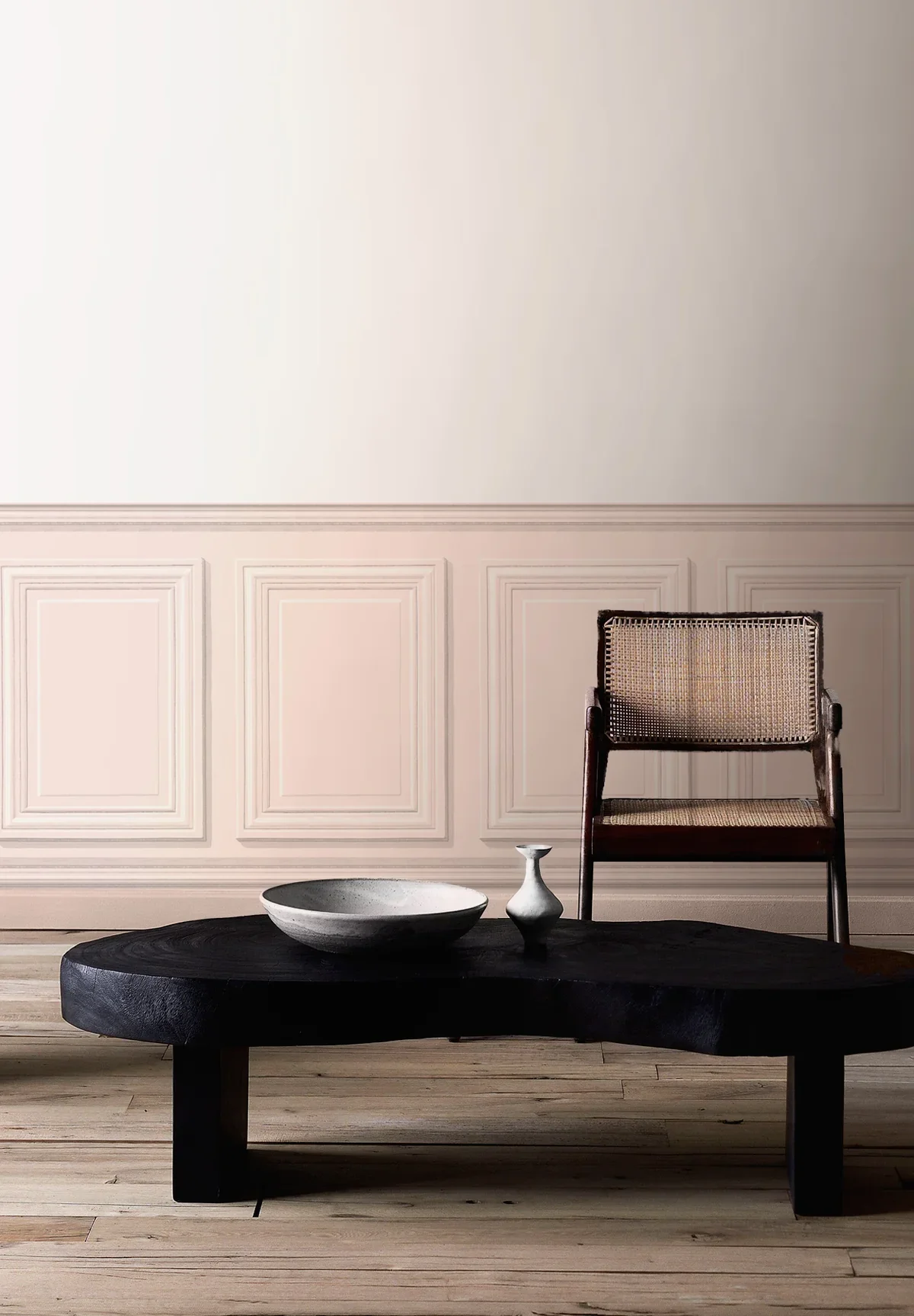 Minimalist room with a dark wooden coffee table, a white ceramic bowl, and a small white decorative vase on it. There is a wooden chair with a cane backrest in the background, against a paneled wall with a soft beige color.