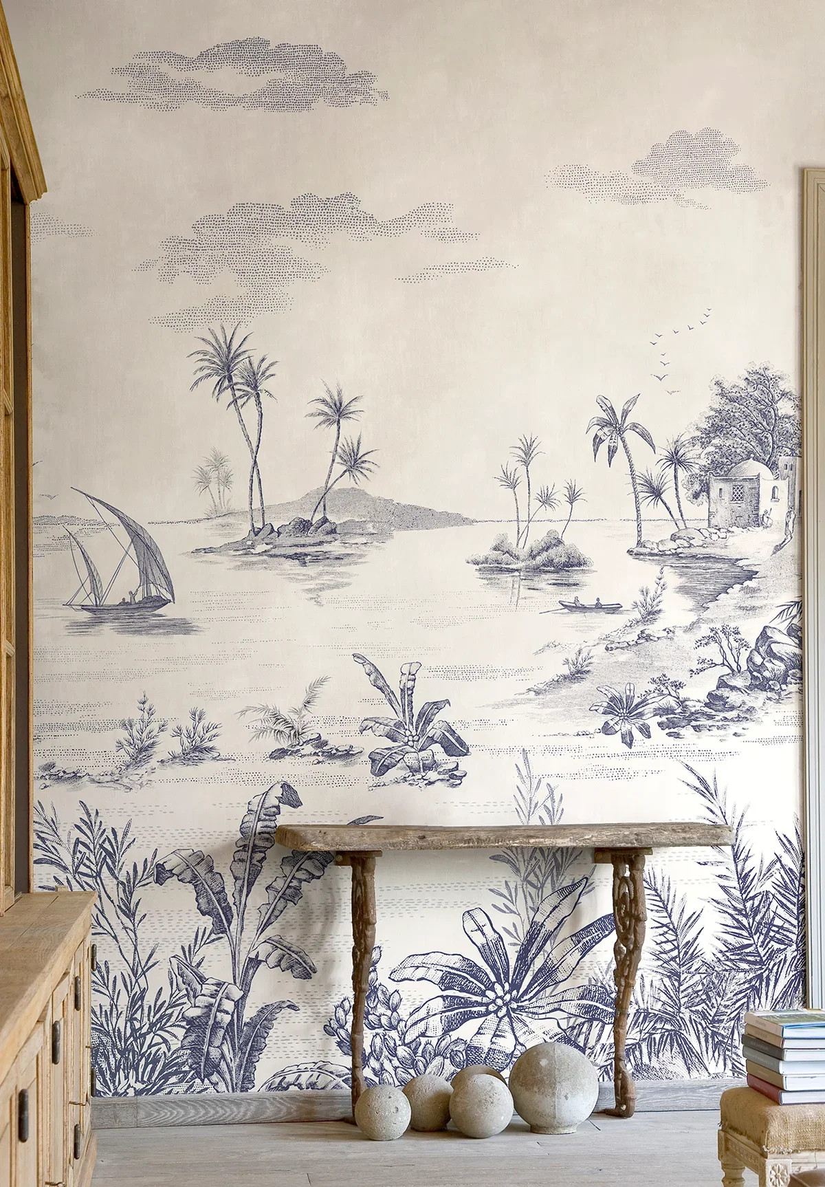 A wall mural depicting a tropical landscape with palm trees, a river, small boats, a sailboat, and a house surrounded by trees in a monochromatic blue and white style. A rustic wooden table and rocks are in front of the mural.