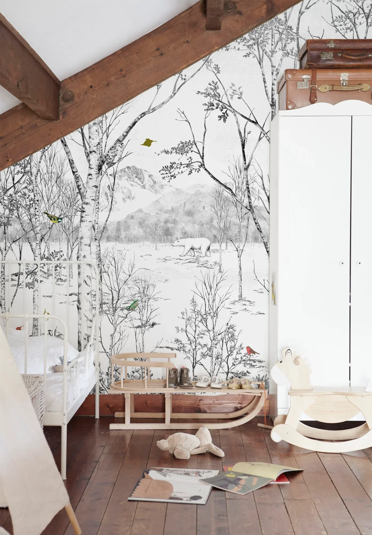 A child's bedroom with winter woodland mural on wall, including trees, mountain, bear, and birds. Bed with white bedding, plush toy on wooden floor, and a rocking horse. Old-style travel trunks on top of white dresser.