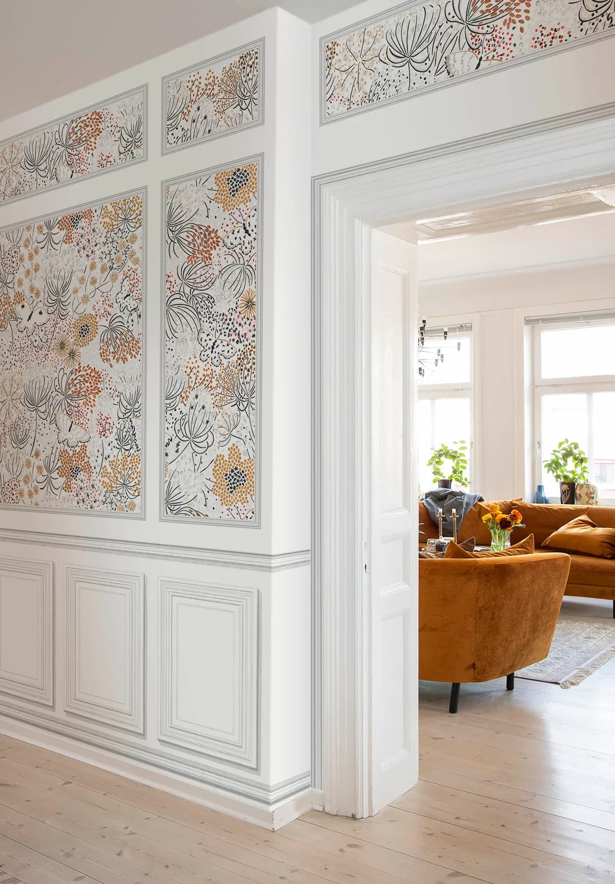 Interior view of a room with decorated walls, a white door frame, and a living room with orange velvet sofas, large windows, potted plants, and a small table with a flower arrangement.