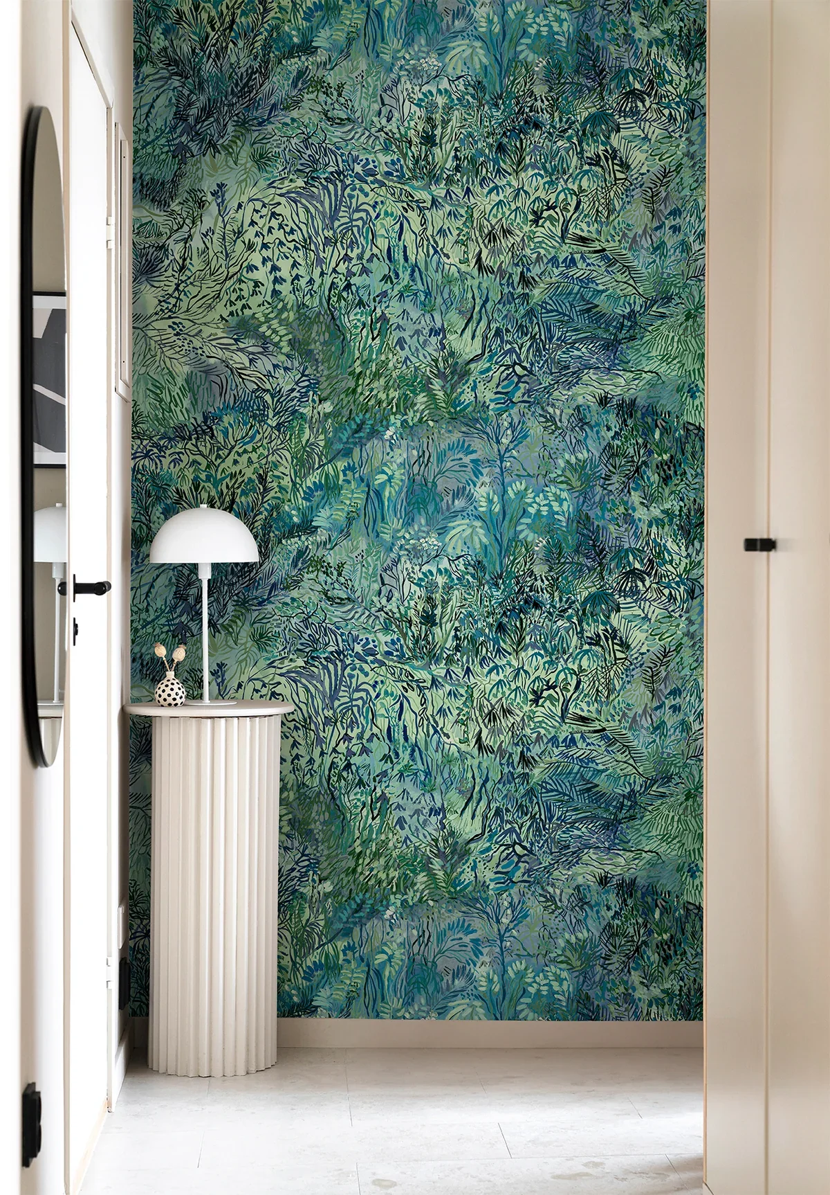 A vibrant indoor wall decorated with a leafy, tropical wallpaper in shades of green and blue. There is a small white pedestal table with vertical ridges on the left side, topped with a white, dome-shaped table lamp and a decorative object with polka 