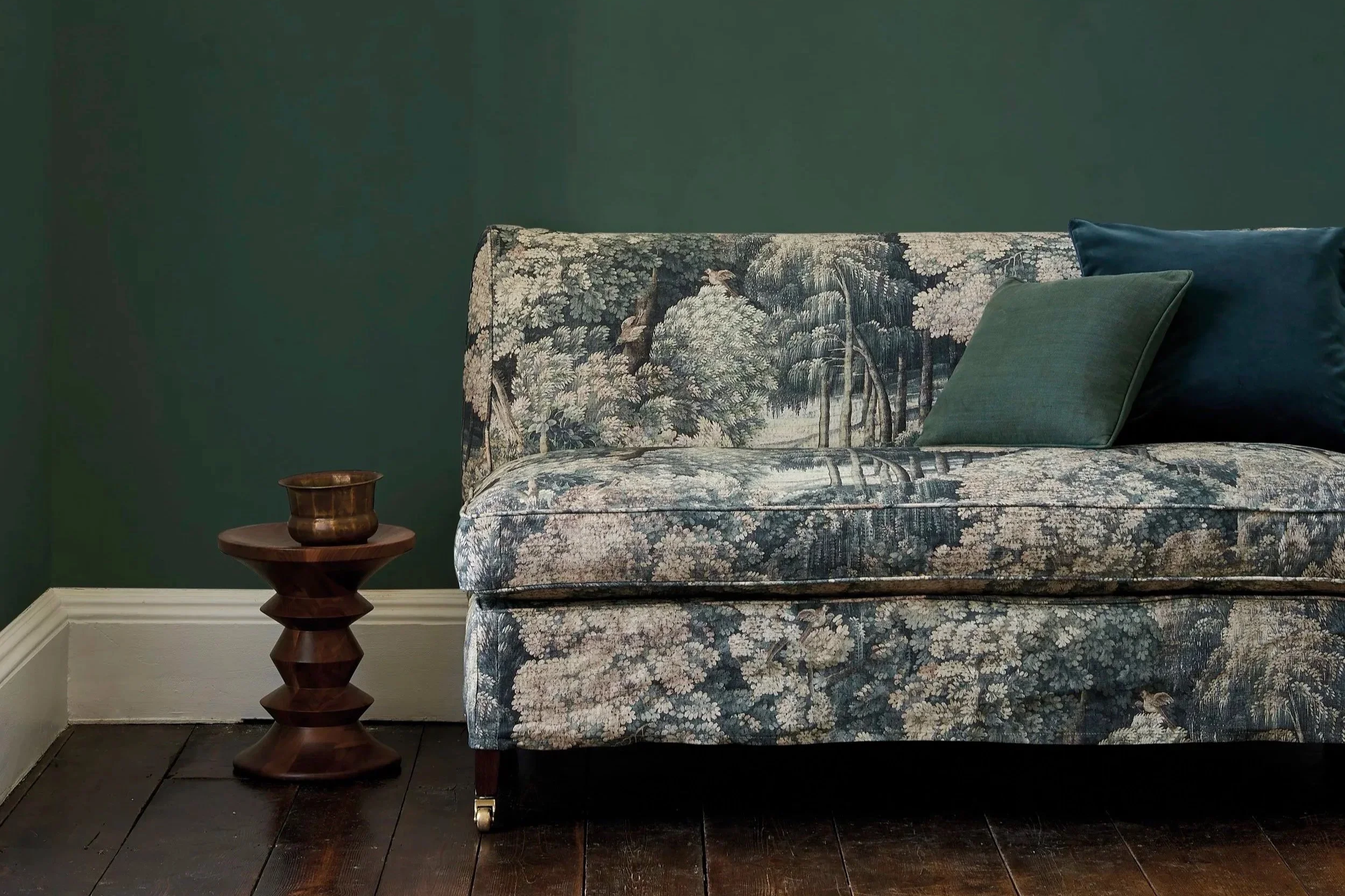 A vintage-style sofa with a floral and woodland tapestry pattern, two pillows (one green and one blue), beside a small wooden side table with a copper-colored decorative vessel on it, against a dark green wall. The sofa has wooden legs with brass casters and rests on dark wood flooring.