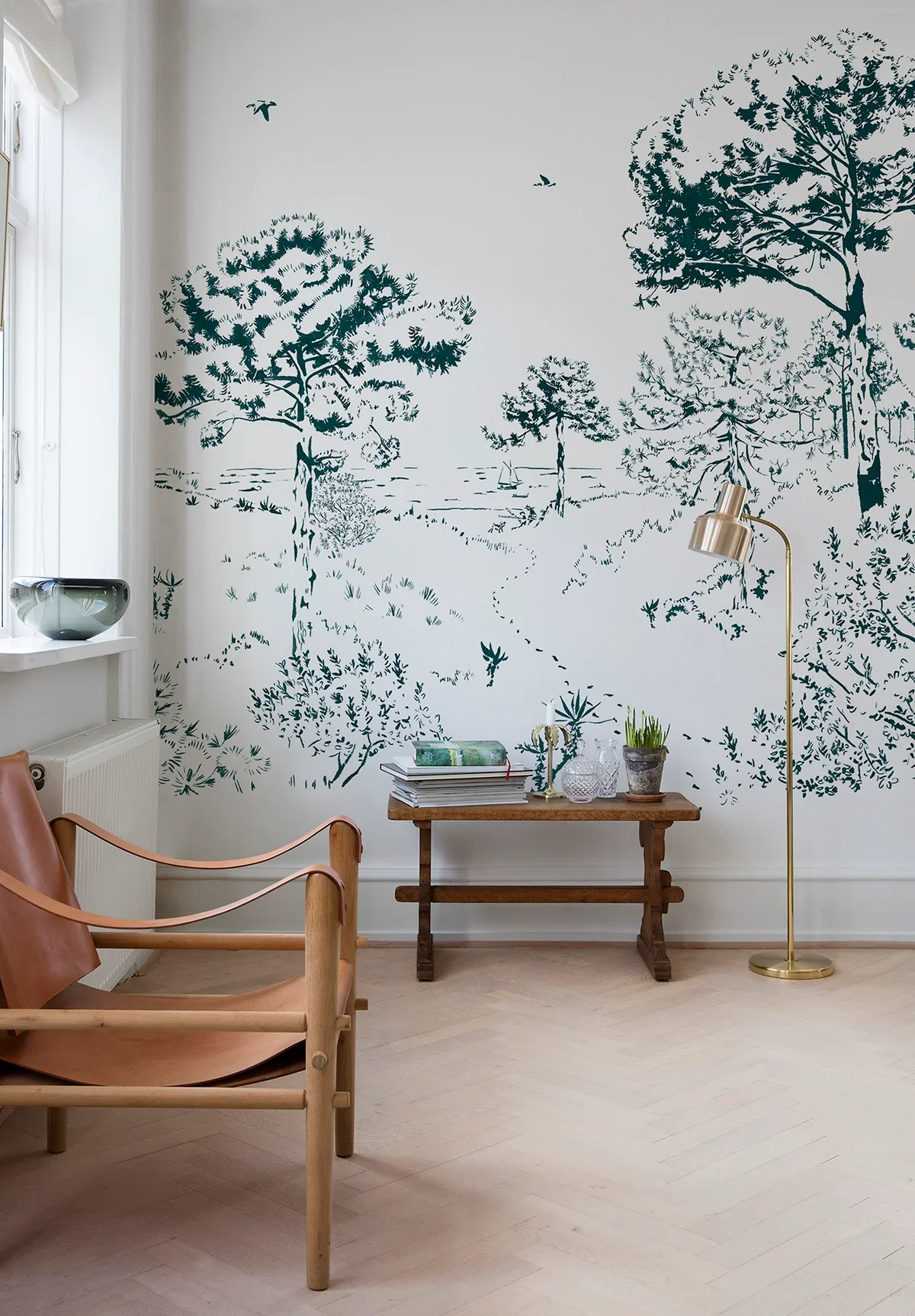 Interior of a room with a white wall decorated with green silhouette trees and birds. A wooden chair with leather seat, a small wooden table with books and plants, a tall floor lamp, and a window with white curtains.