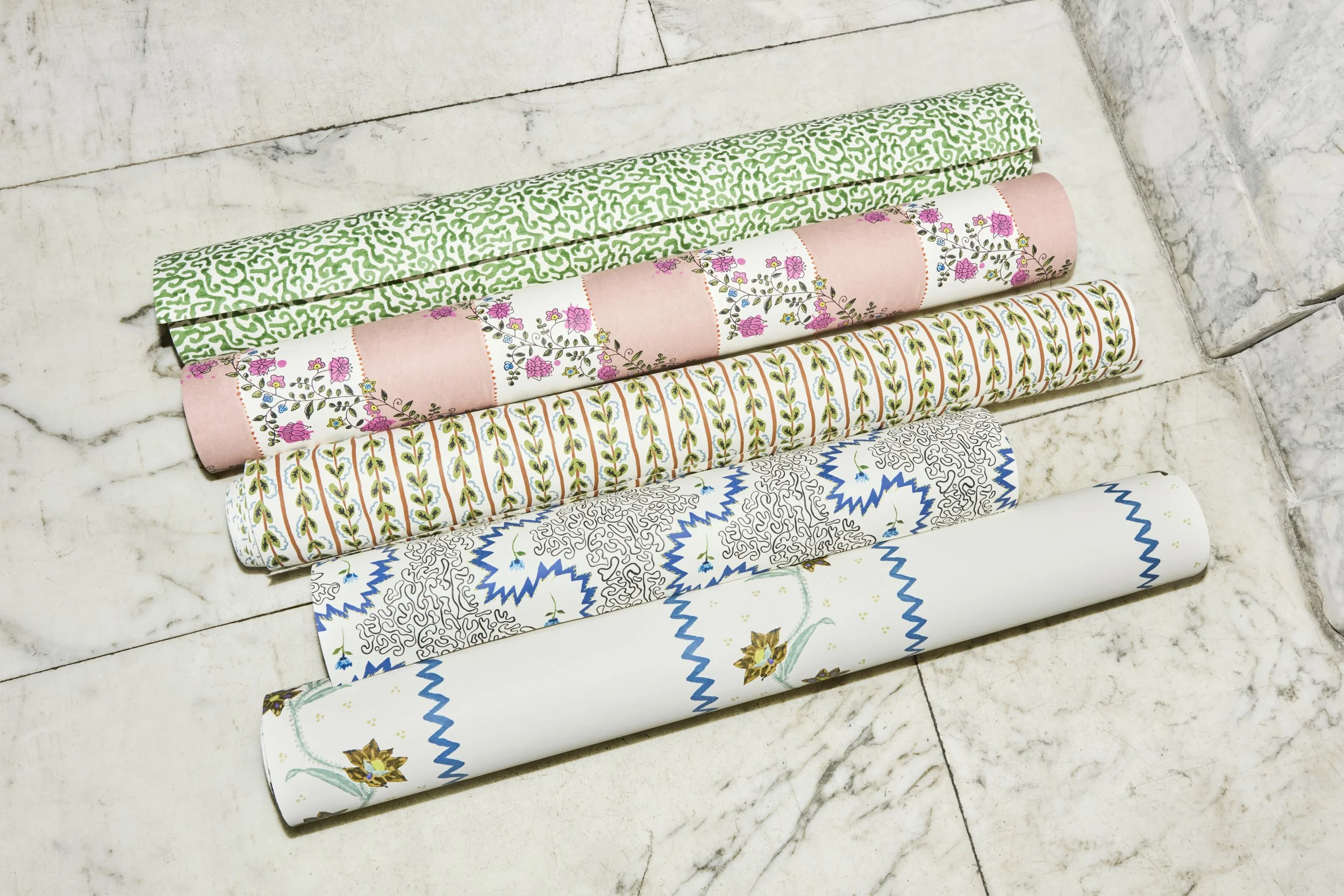 Five rolls of decorative patterned wallpaper stacked on a marble surface.
