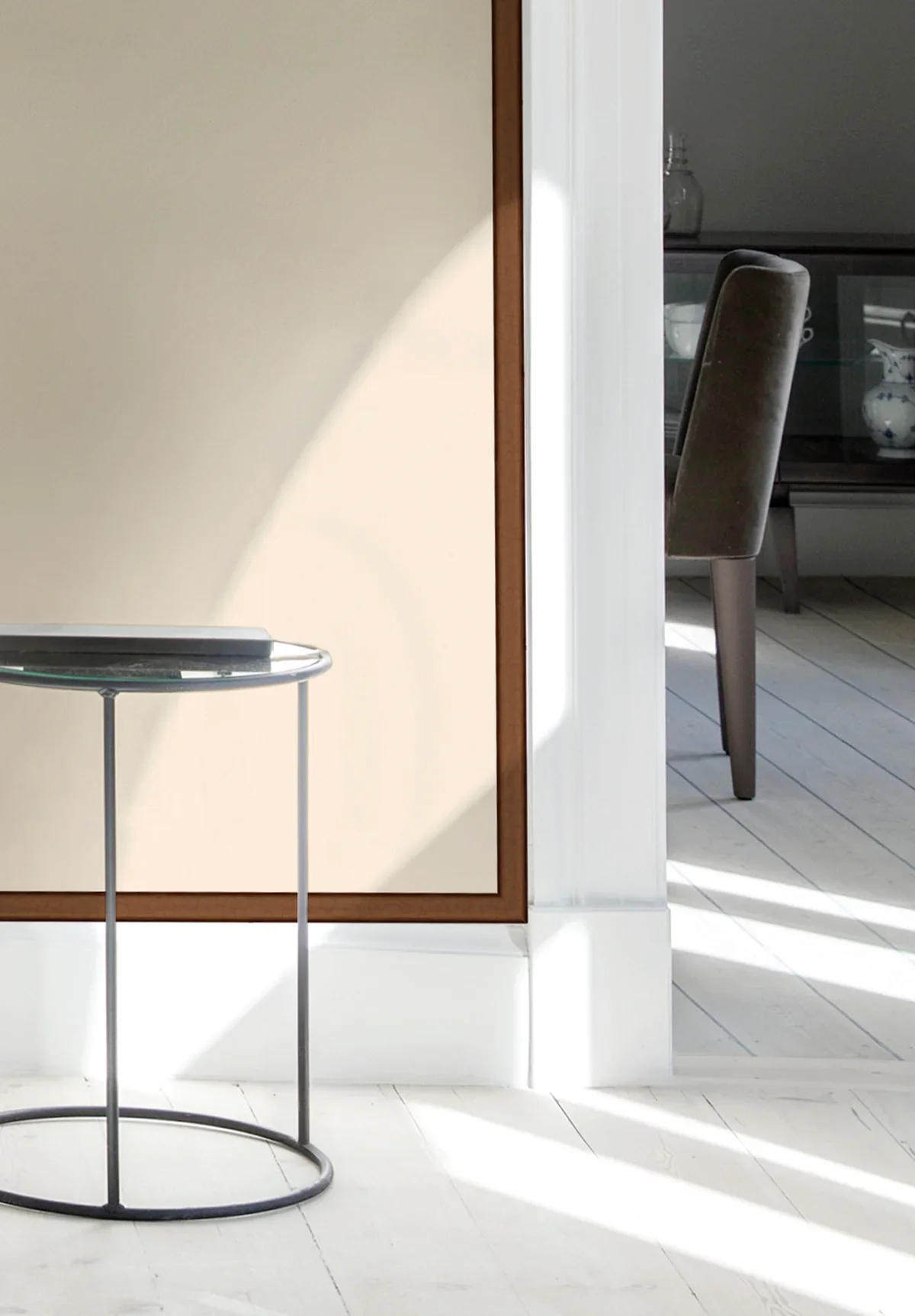Part of a modern interior with white walls, a small round glass-topped side table, and a view of a dining area with a dark upholstered chair and a glass-front cabinet with dishes.