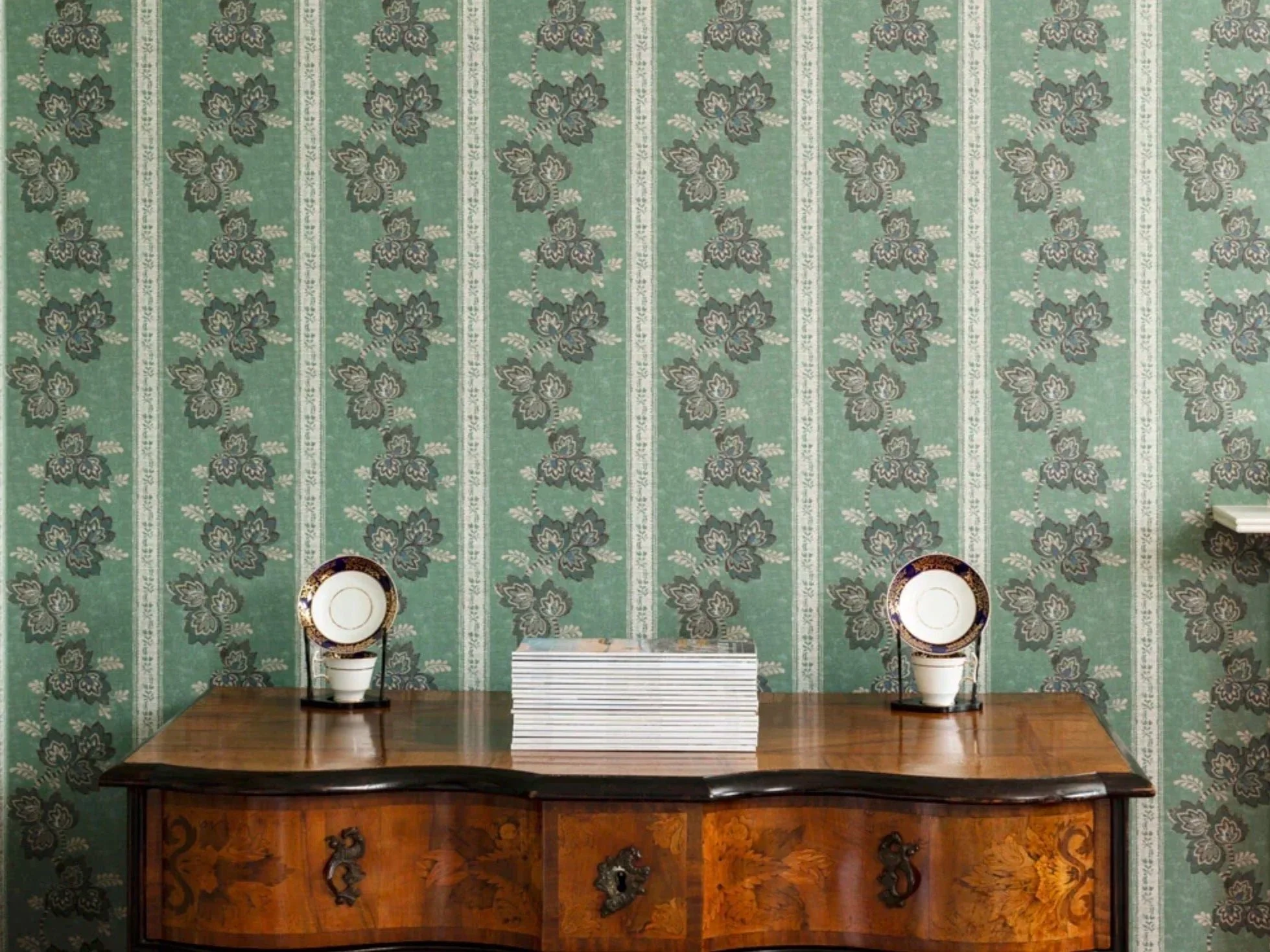 A wooden sideboard with a stack of magazines on top, flanked by two decorative plates on stands against a green floral-patterned wallpaper.