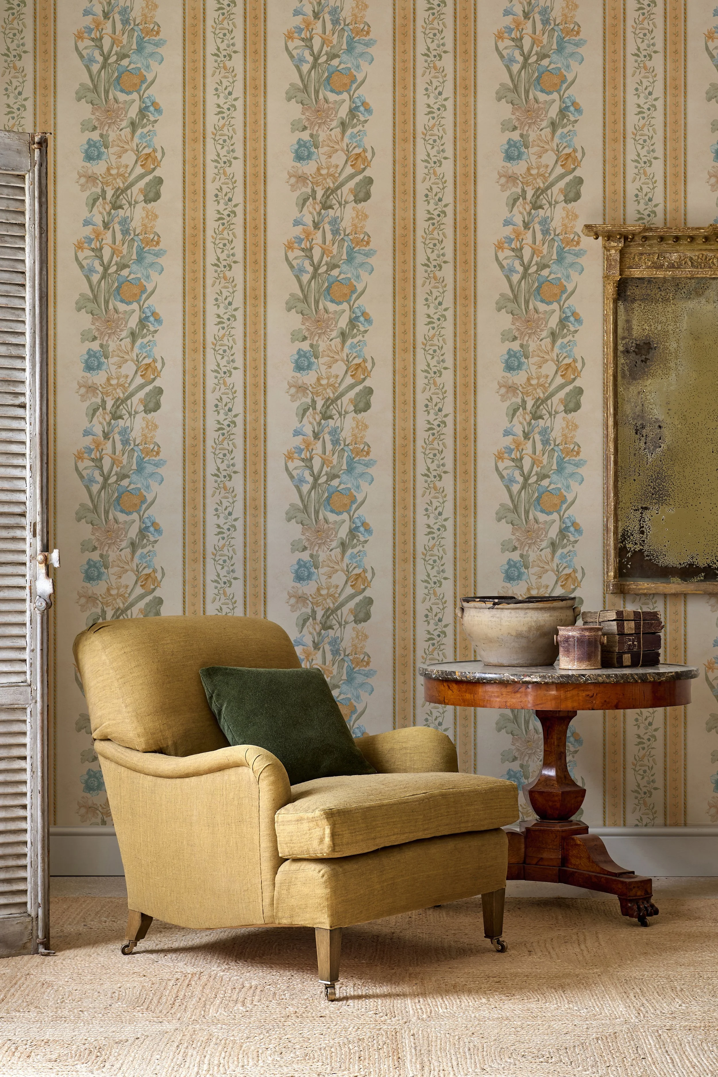 Interior of a vintage living room with cream-colored wallpaper featuring a floral pattern, a mustard yellow armchair with a dark green pillow, a round wooden side table with antique books and a large ceramic bowl, and a distressed gilded mirror frame on the wall.