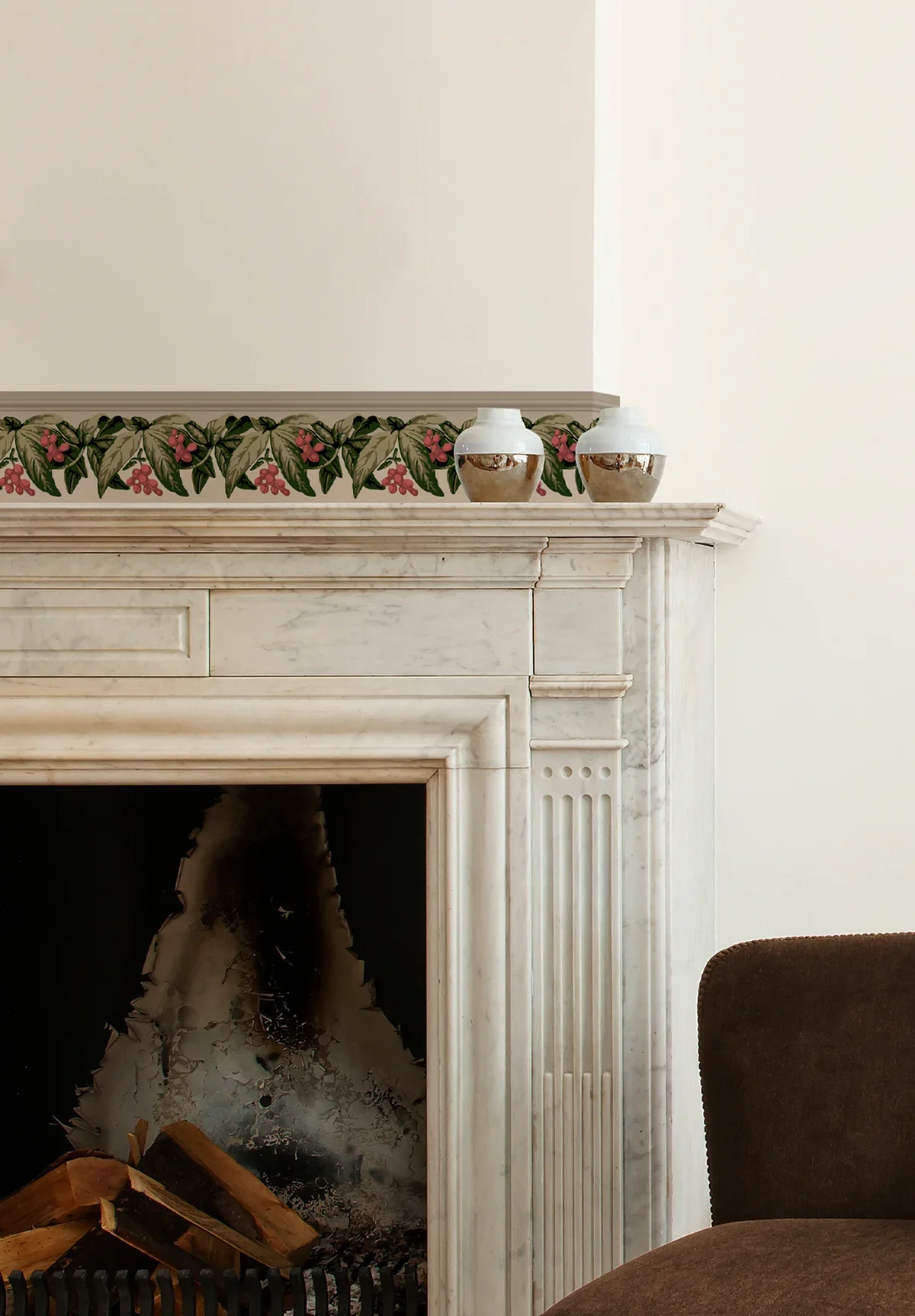 A white marble fireplace with decorative carvings and a floral border at the top, topped with two ceramic vases. The fireplace opening shows a fire with logs inside, and part of a brown upholstered chair is visible on the right side.