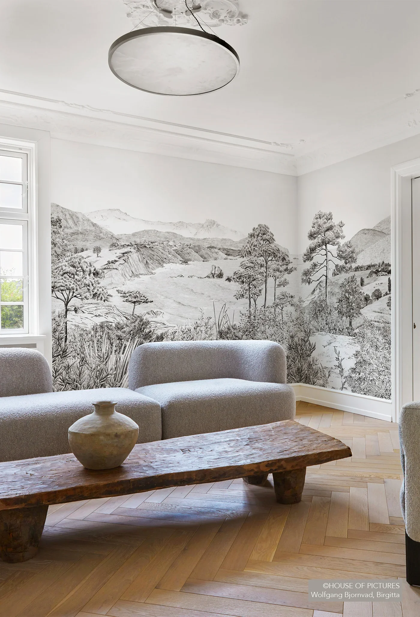A living room with a mural of a black-and-white landscape depicting mountains, trees, and a river on the wall behind a gray sofa and a rustic wooden coffee table with a ceramic vase.