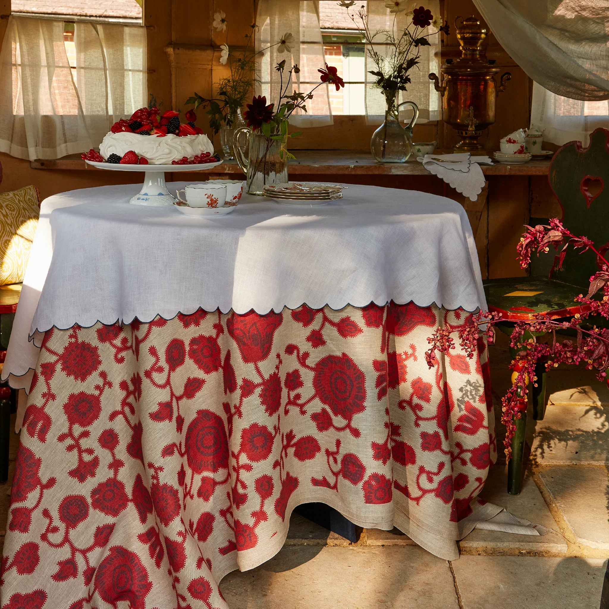 A table set with a white tablecloth, a cake topped with berries, and several flower vases with flowers. In the background, there is a wooden window with sheer curtains and various antique kitchen items.