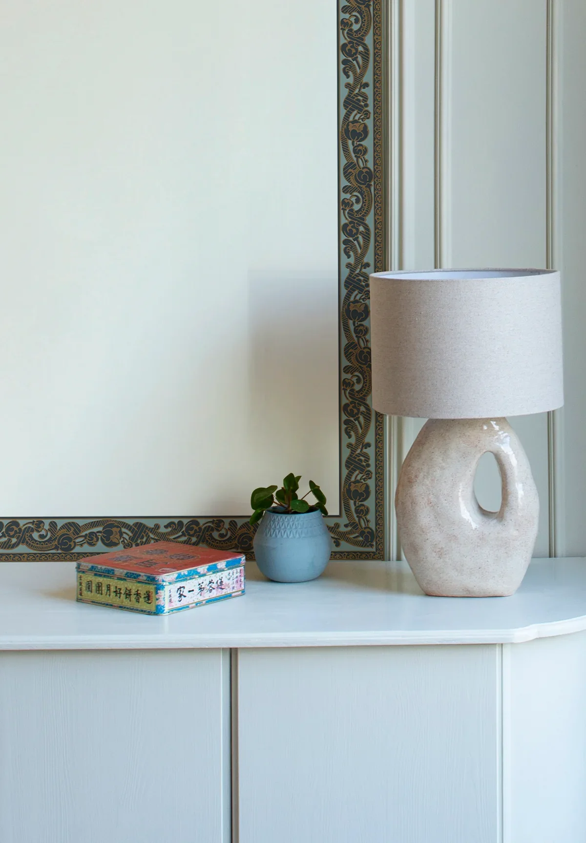 Decorative table with a ceramic table lamp, a small potted plant, and a box with Chinese characters, against a white wall and ornate framed mirror.
