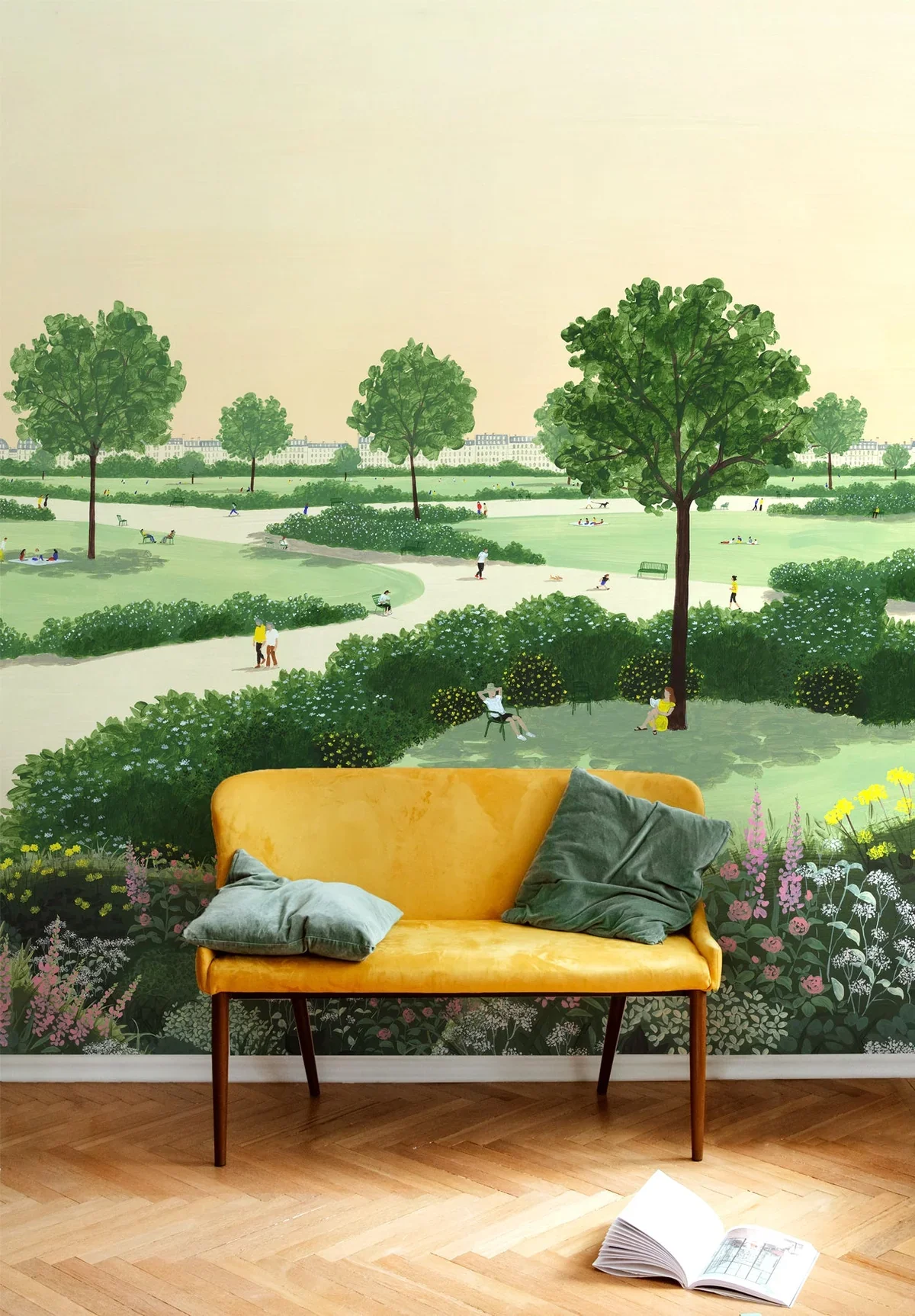 Interior wall with mural of a park scene featuring trees, bushes, pathways, and people enjoying the outdoors. There are two green pillows on a yellow sofa and an open book on the wooden floor.