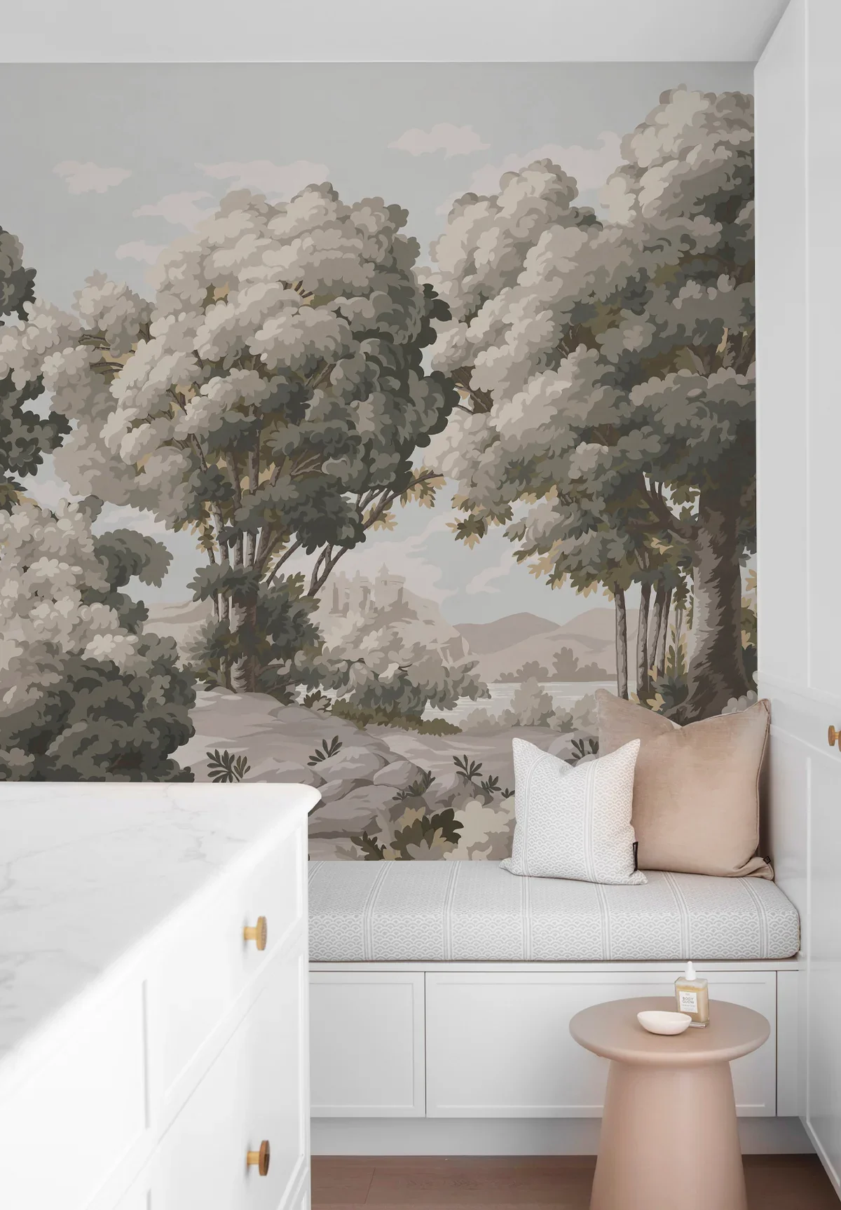 A cozy sitting nook with a white built-in bench, decorated with beige and white pillows, beneath a mural of a forest landscape with trees, rocks, and distant hills. A small beige side table holds a bottle of lotion and a small dish.