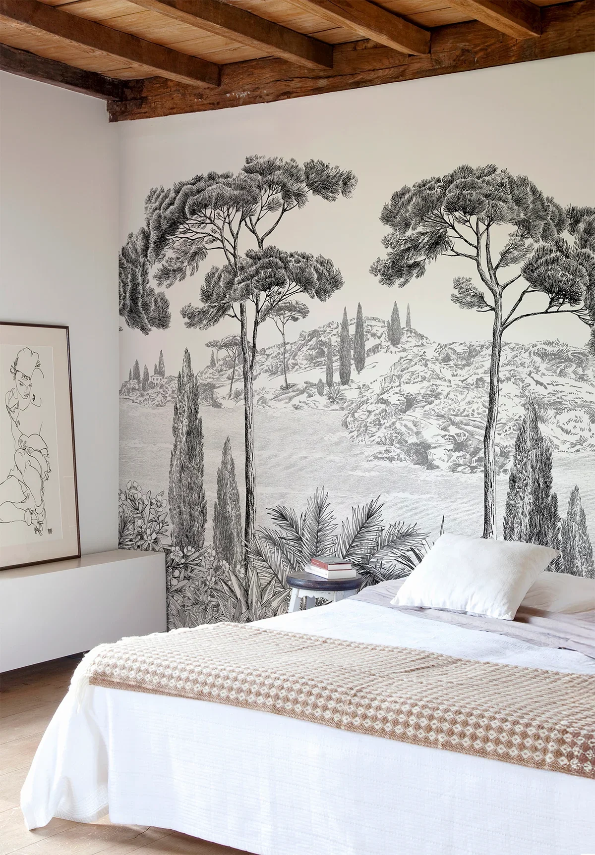 A bedroom with a mural of trees, mountains, and a body of water behind a bed with white bedding and a beige blanket.