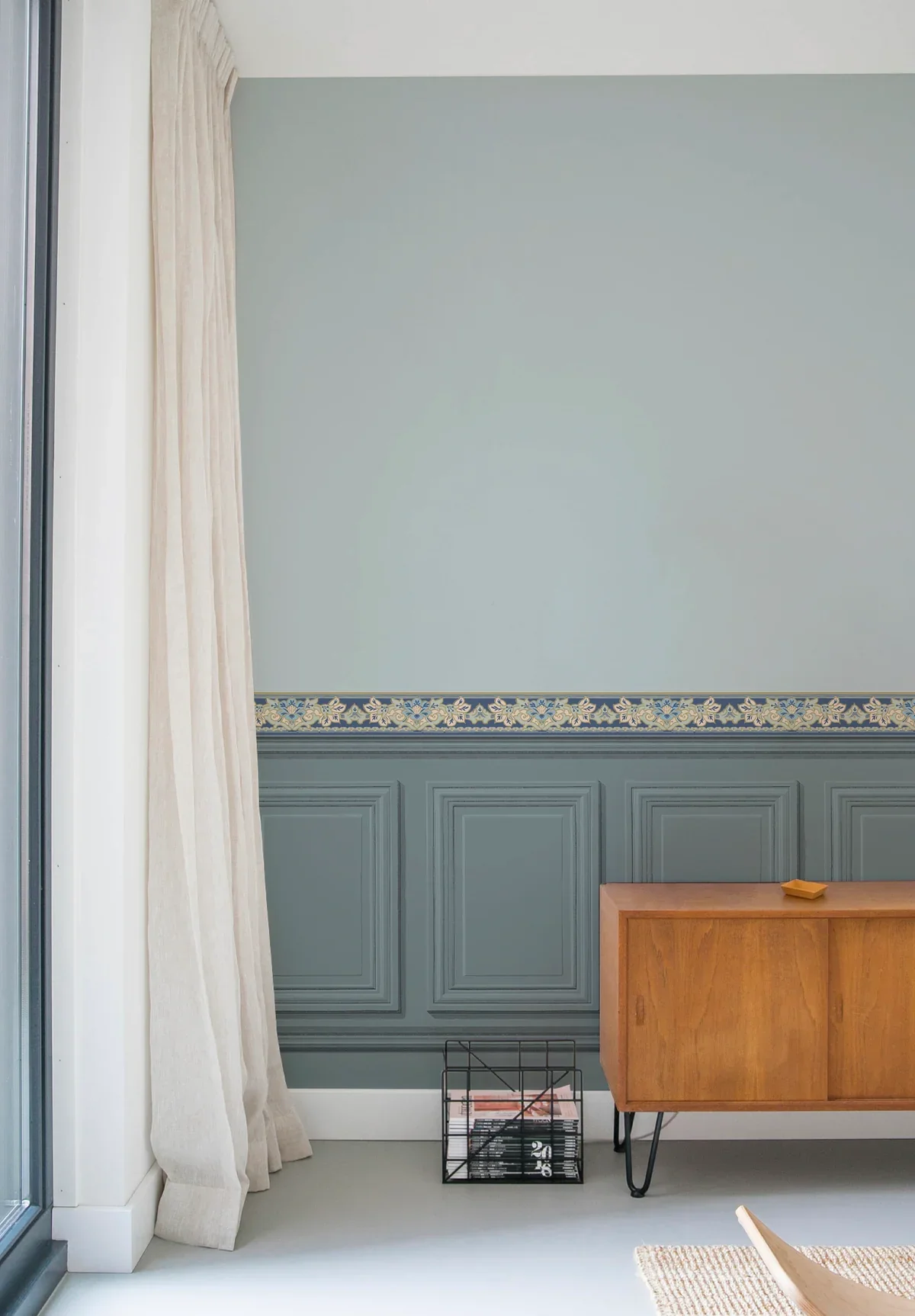 Interior of a room with a pale blue wall, decorative blue and gold border, a wooden sideboard, a black wire magazine holder with magazines, and a beige curtain next to a glass sliding door.