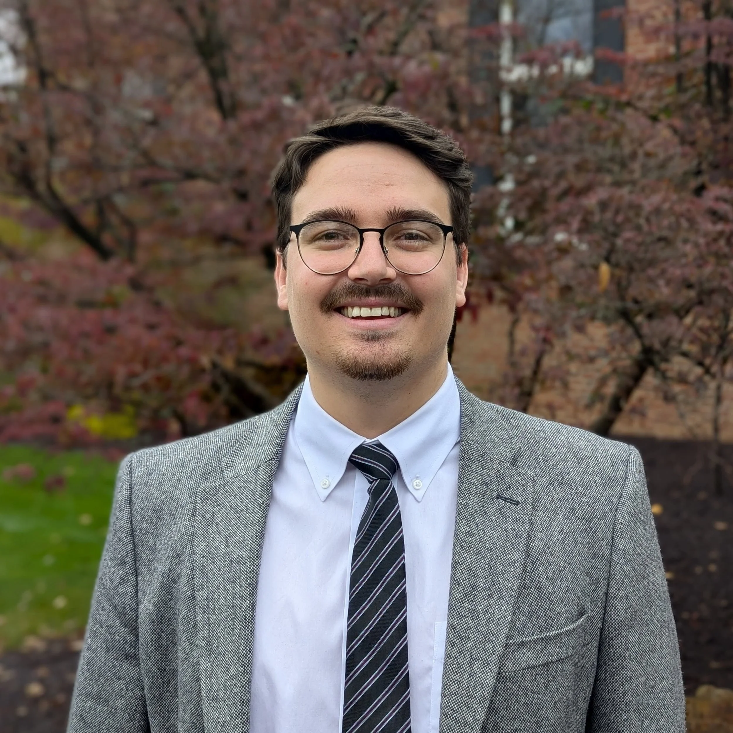 Matthew Ball in black frame glasses wearing a gray jacket and black and gray striped tie.