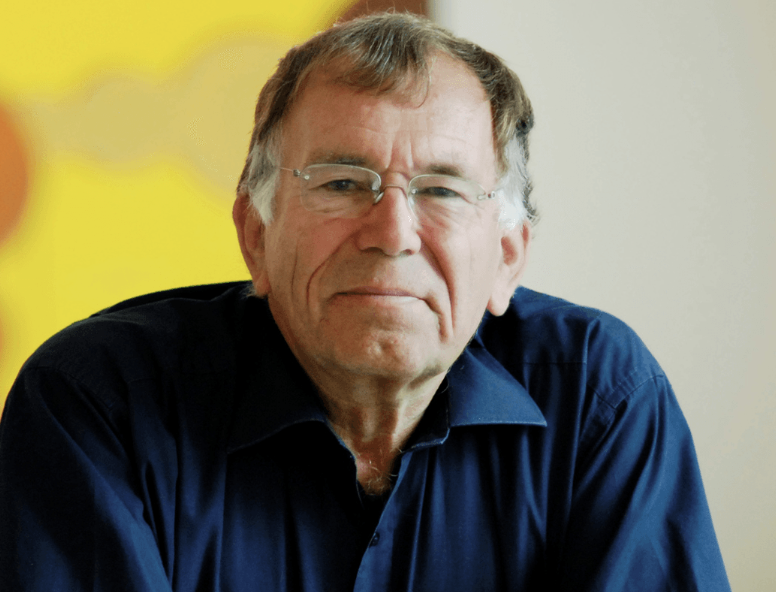 A middle-aged man with glasses and gray hair wearing a dark blue shirt, sitting indoors with a yellow wall in the background.
