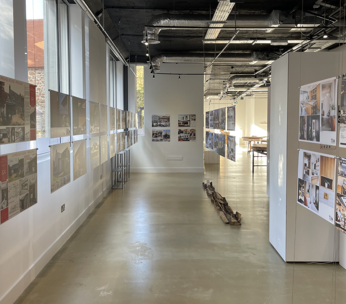 An art gallery or exhibition space with photographs and images displayed on white walls and panels, featuring interior and architectural designs, with large windows letting in natural light and a polished concrete floor.