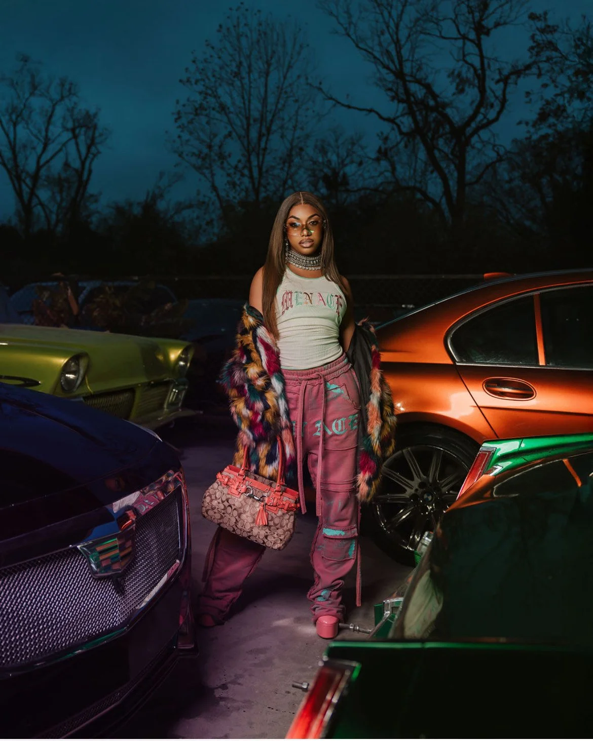 A fashionable woman standing among luxury cars at night, wearing a fur coat, sunglasses, and holding a designer handbag.
