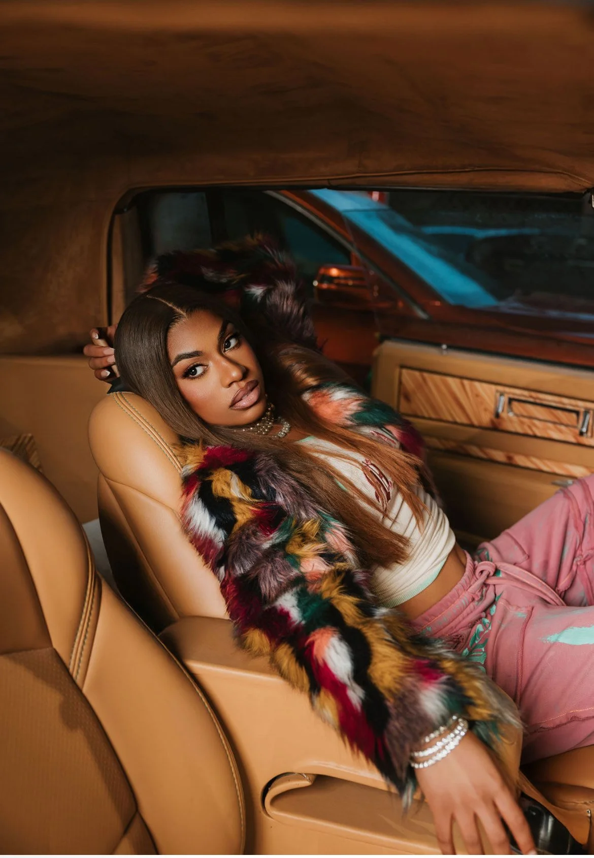 A woman with long brown hair and makeup leaning back in a tan leather car seat, wearing a colorful fur jacket, a beige crop top, pink pants, and jewelry, inside a vintage car.
