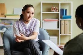 A woman sitting in a chair during a therapy or counseling session, talking to another person holding a notepad.