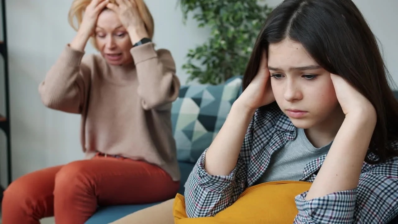 An older woman and a young girl sitting on a couch holding their heads, both appearing stressed or upset.