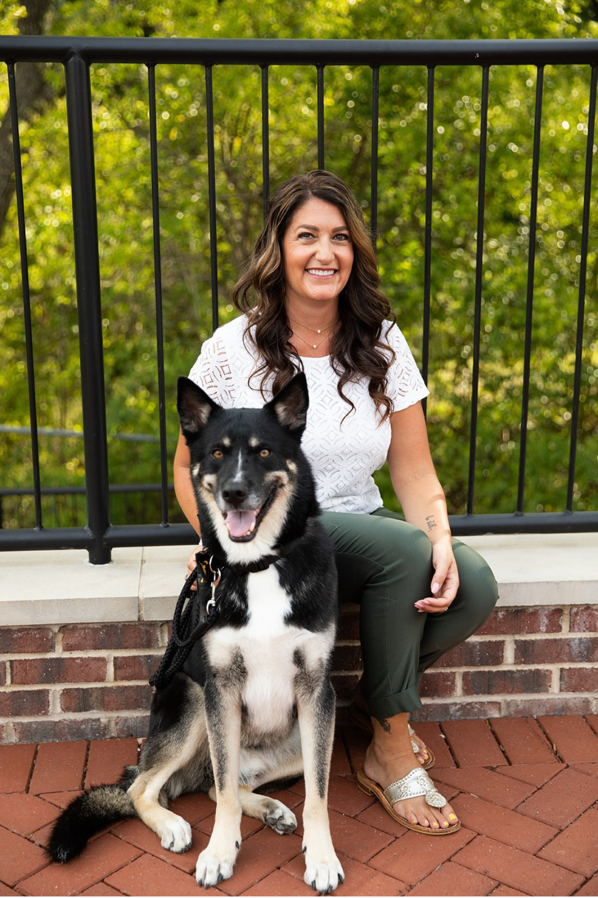 Dr. Tonyia Behrendt with long wavy brown hair smiling, wearing a white patterned top and green pants, sitting on a brick ledge next to a black and white husky dog with amber eyes outdoors surrounded by green trees.