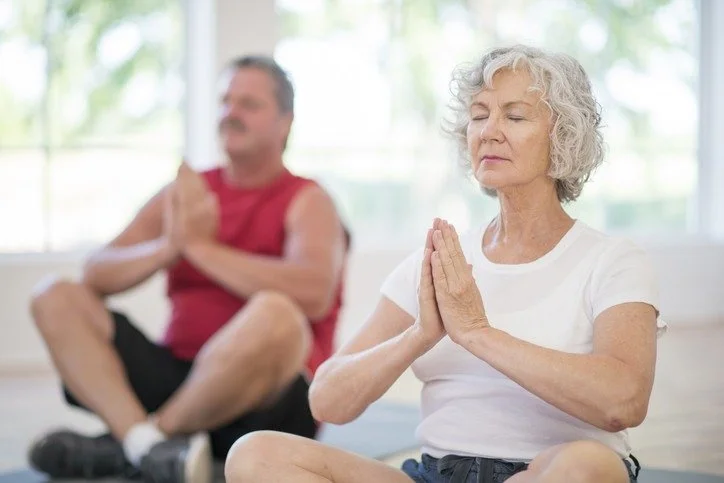 An elderly woman and a middle-aged man practicing yoga or meditation indoors. The woman is sitting cross-legged with hands in a prayer position, eyes closed. The man is sitting cross-legged with hands in a prayer position, eyes closed.