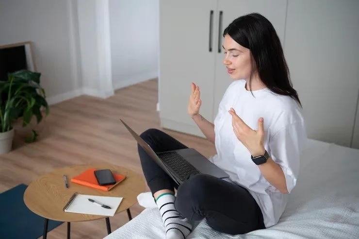 Young woman sitting on her bed, waving at her laptop with a smile, in a bright room with a small table holding a phone, notepad, and pen nearby.