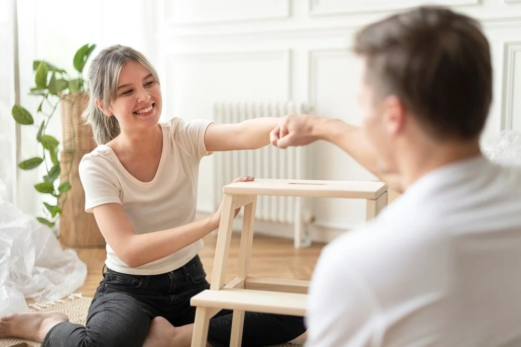 A young girl with blonde hair smiling and reaching out to fist bump a man, sitting on the floor in a bright room, with a small wooden table between them.