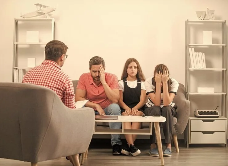 A family of four sitting on a couch during a therapy session with a counselor, showing signs of distress, with a therapist taking notes.