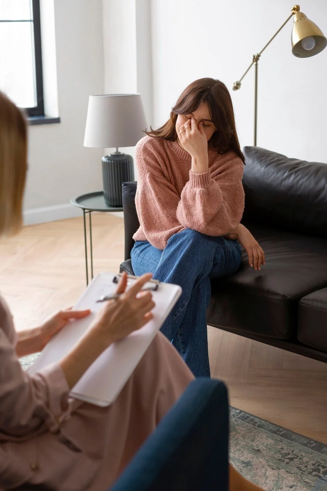 A woman with brown hair wearing a pink sweater and blue jeans sitting on a black leather couch, holding her face with her hand, appearing upset, during a therapy session with a therapist taking notes.