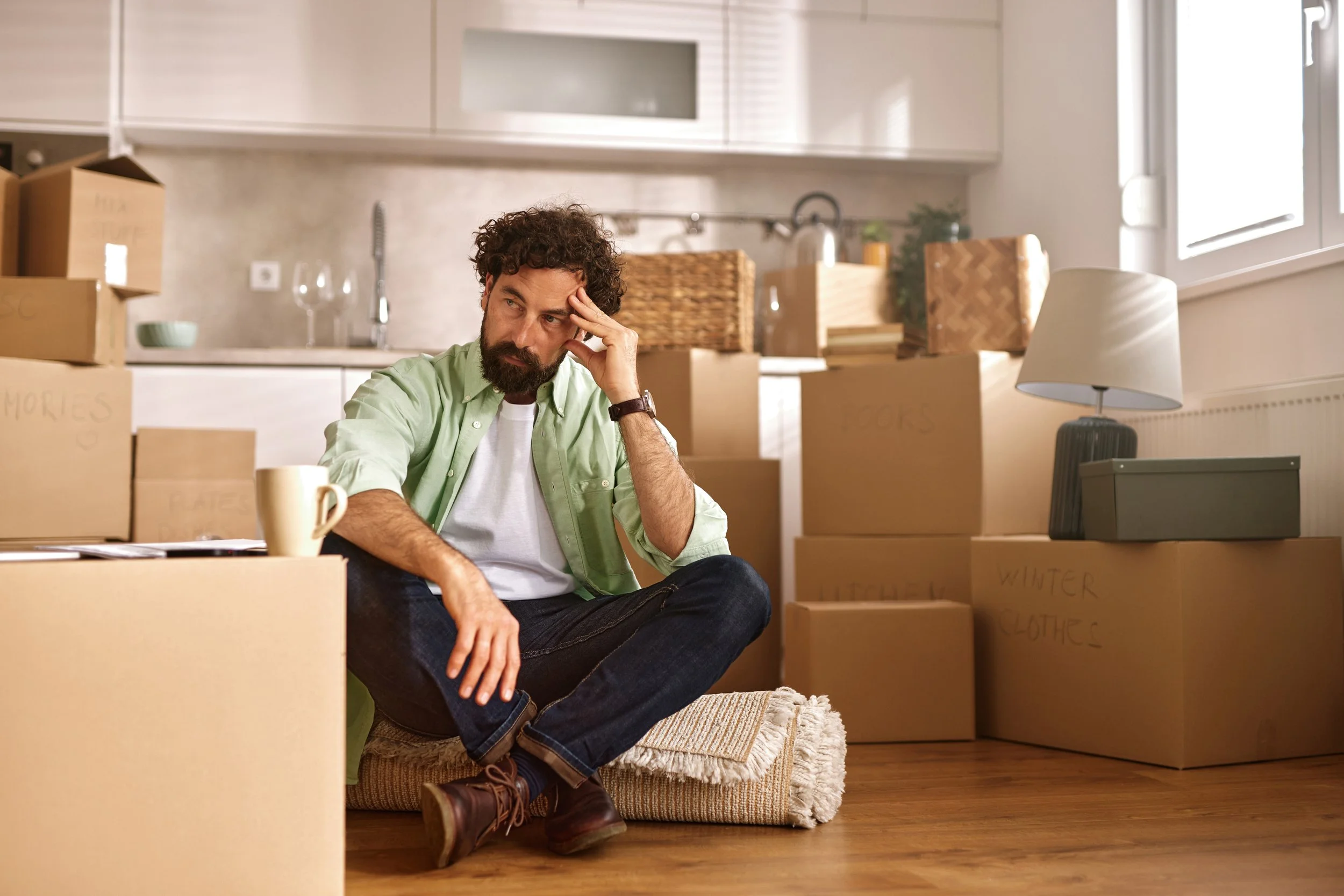 A man with a beard and curly hair sits on a rug on the floor, surrounded by cardboard moving boxes, in a kitchen. He appears to be stressed or tired, with his hand on his forehead and a concerned expression.