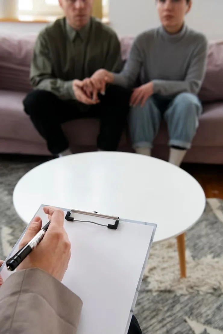 Two people sitting on a sofa with a person in the foreground writing on a clipboard, appearing to conduct an interview or assessment.