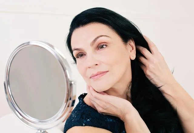 A woman with black hair looking at herself in a mirror, touching her hair and chin with a soft smile.