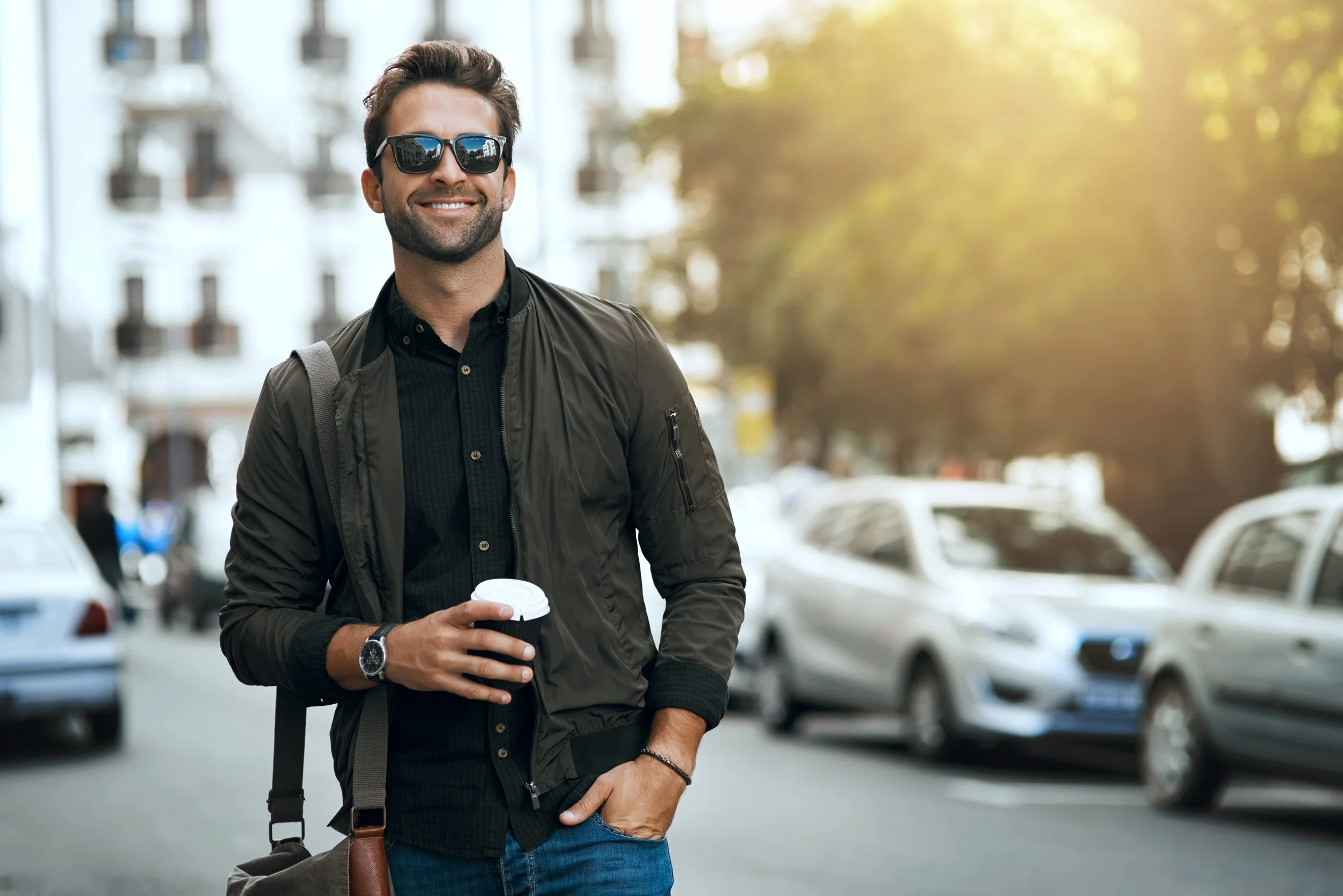 Smiling man in sunglasses holding a coffee cup walking outdoors in an urban area during daytime