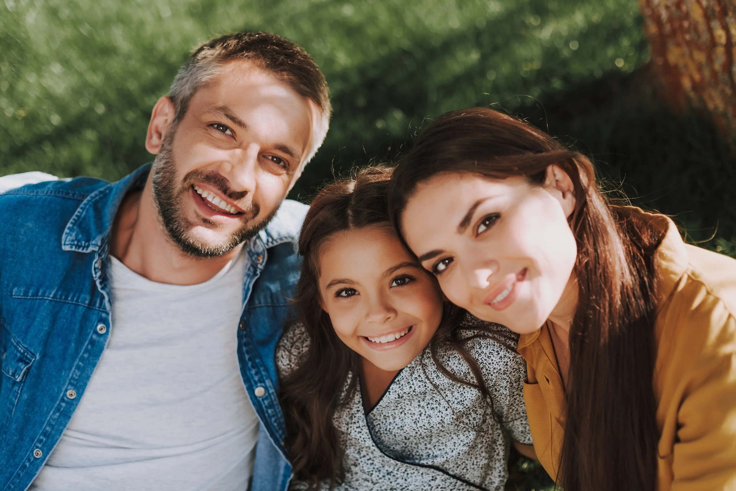 Family of three taking a selfie outdoors on a sunny day, smiling, with green grass and sunlight in the background.