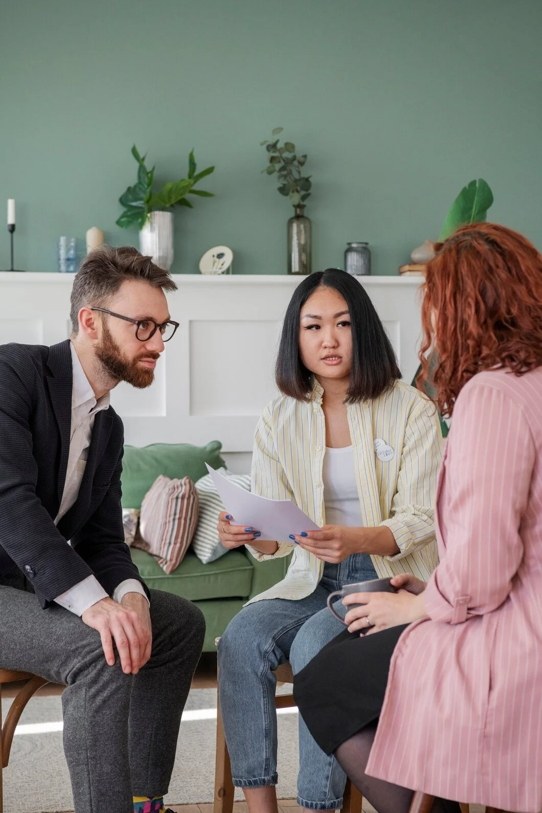 Three young adults in a meeting or discussion in a cozy, well-decorated living room, with two women and one man, two of them holding papers and a mug.