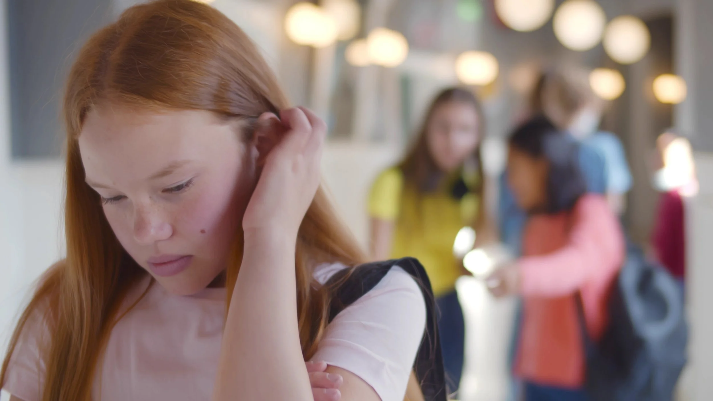 A young girl with red hair is touching her ear and looking down, with a blurred background of other children in a school or library setting.