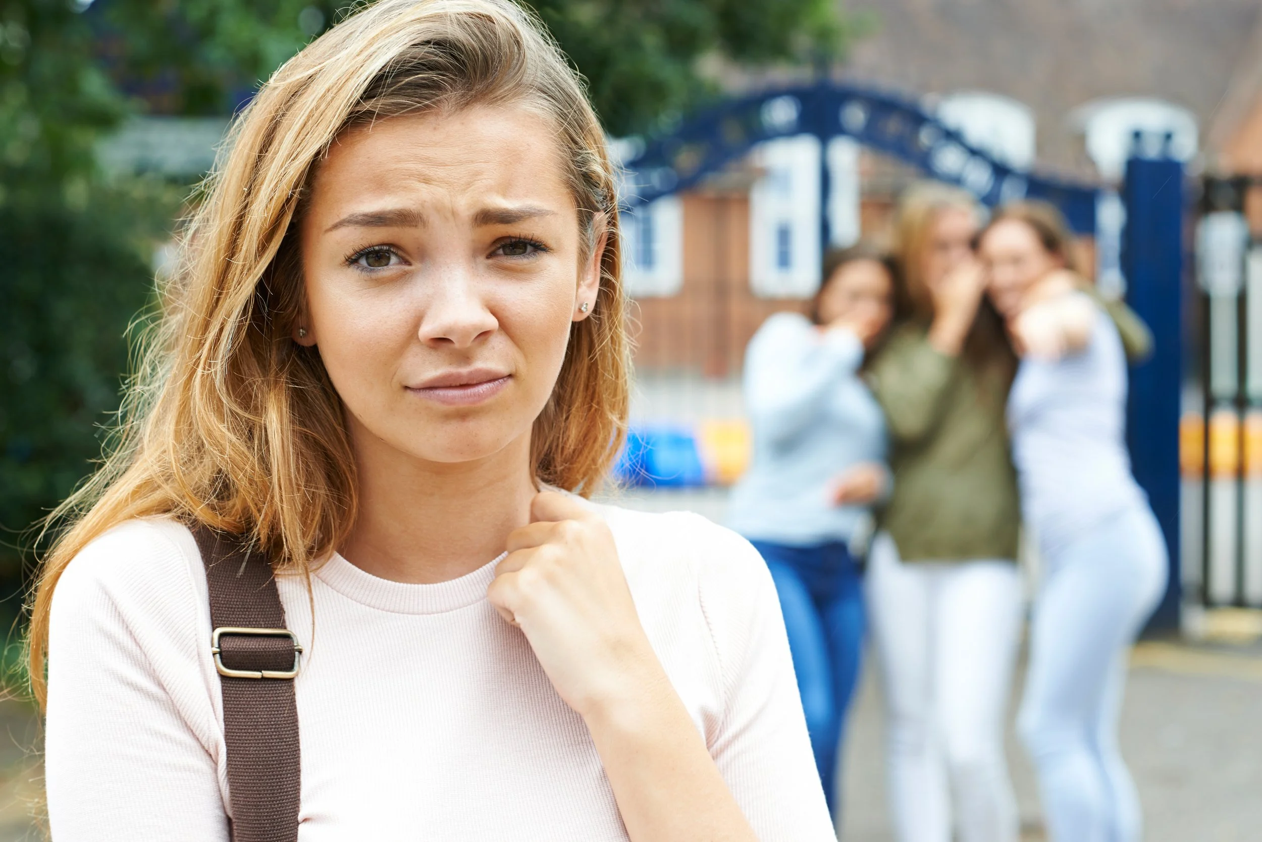 A young woman with long blonde hair, wearing a white shirt and a backpack, looks upset and uncertain, standing outdoors near a gated area with blurred group of women in the background.
