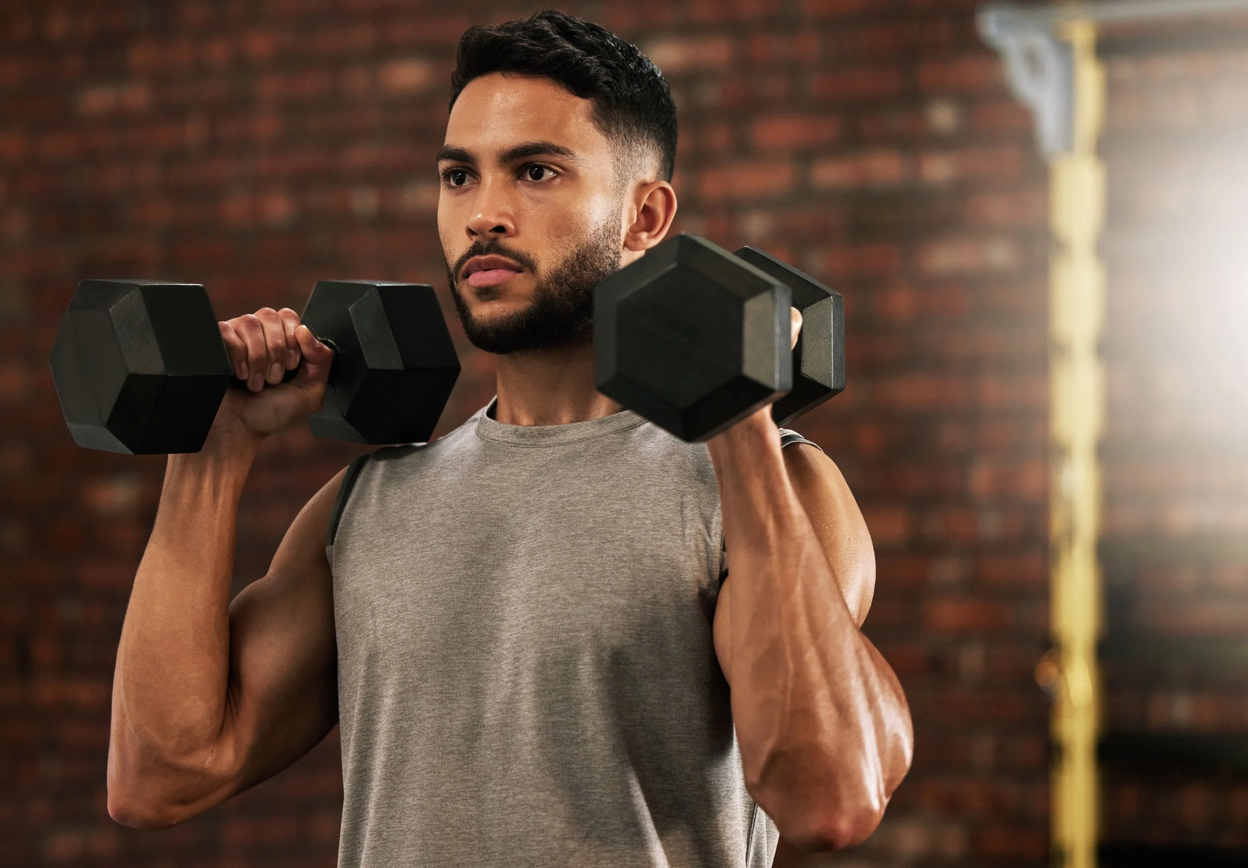 A man in a sleeveless shirt holding a dumbbell in each hand across his shoulders in a gym with a brick wall background.