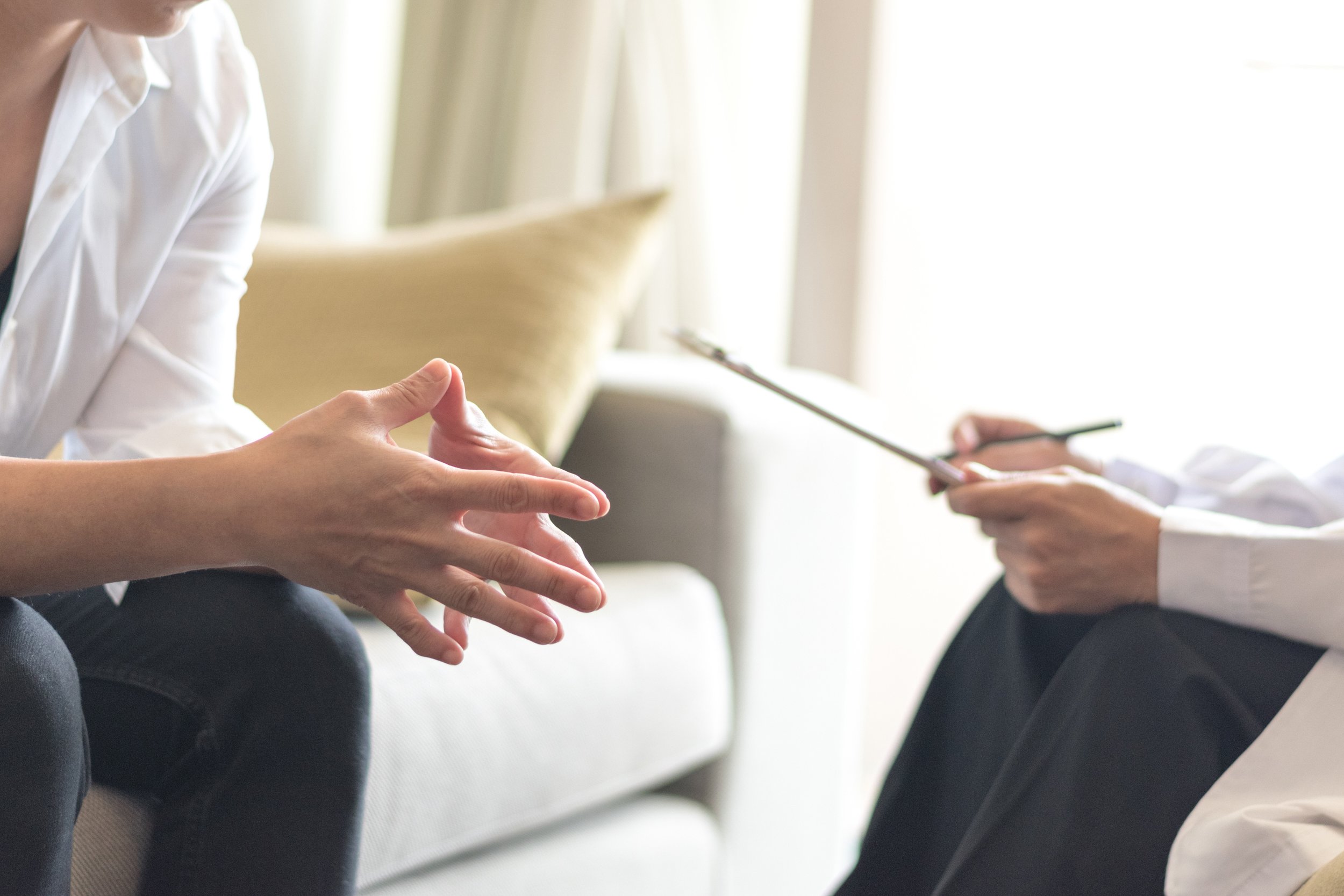 Person sitting on a sofa during a counseling or therapy session, with another person holding a notepad and pen, in a bright room with natural light.