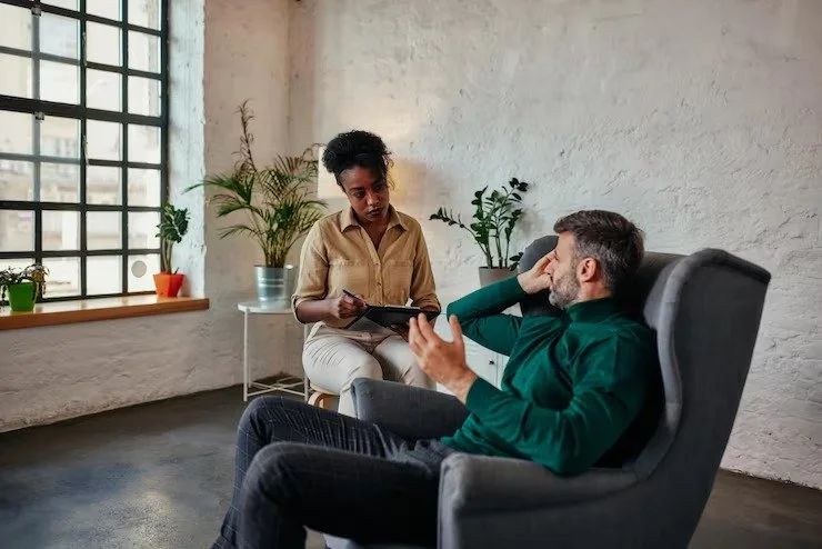 A woman during a therapy session with a man seated in an armchair, holding a tablet, in a room with potted plants and a large window.