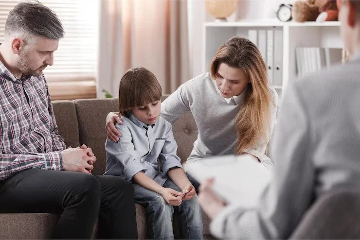 A young boy is sitting on a couch with his parents and a counselor, likely during a therapy session. The boy looks downcast while his parents comfort him, and the counselor holds a clipboard or papers.