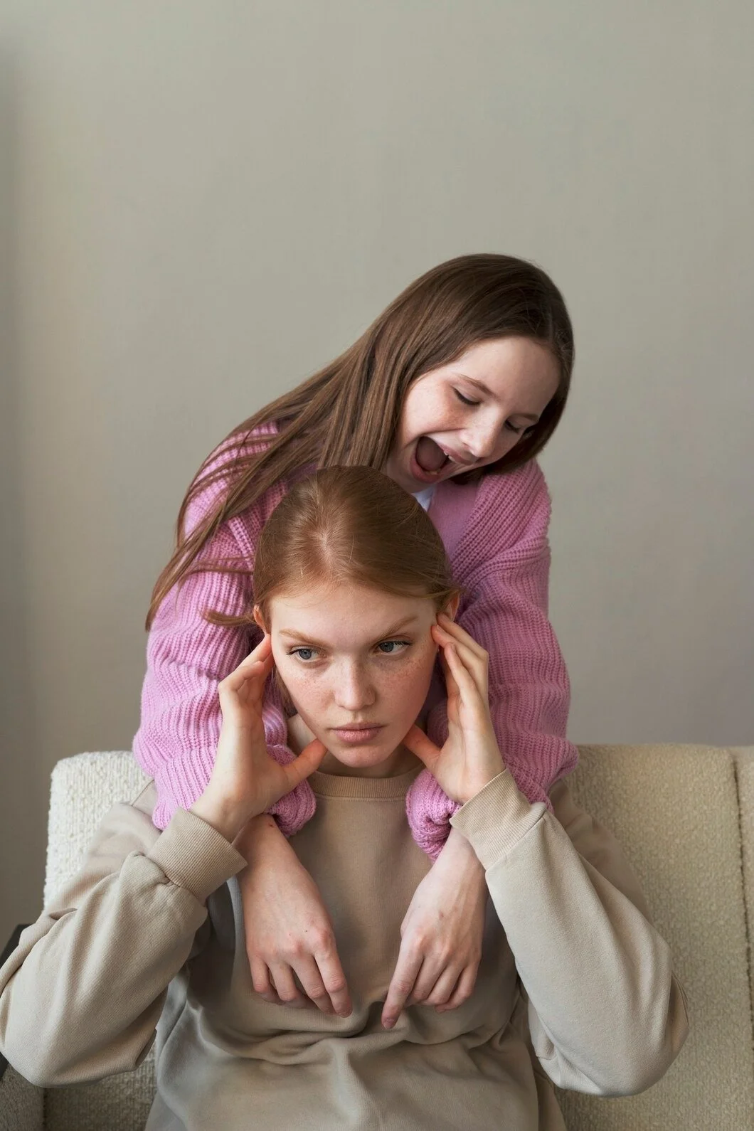 Two young women with red hair, one wearing a pink sweater and the other in a beige top, sitting on a beige couch, with the woman in pink showing a playful or expressive gesture towards the woman in beige.