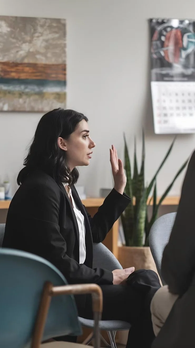 A woman with dark hair, dressed in a black blazer, sitting in a meeting or office environment, raising her right hand.