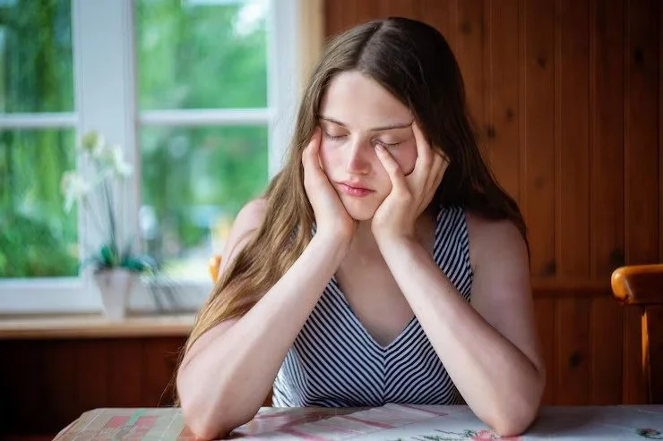 A young woman sitting at a table with her eyes closed, holding her head with both hands, appearing to be upset or tired.